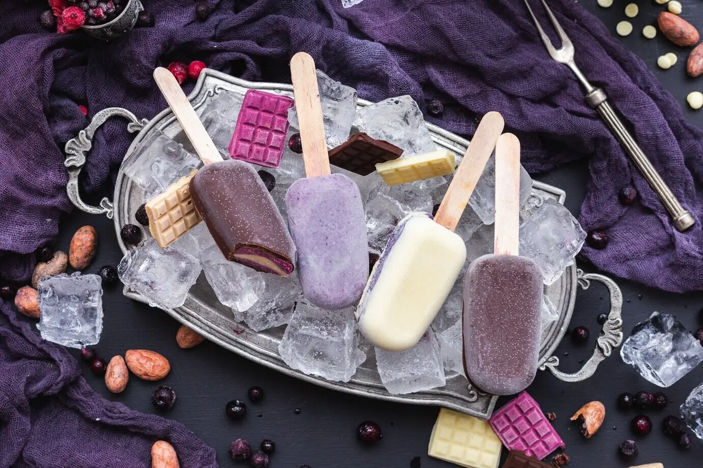 Vertical photo showing homemade vegan ice creams and chocolate bars placed on ice cubes in a metal plate.